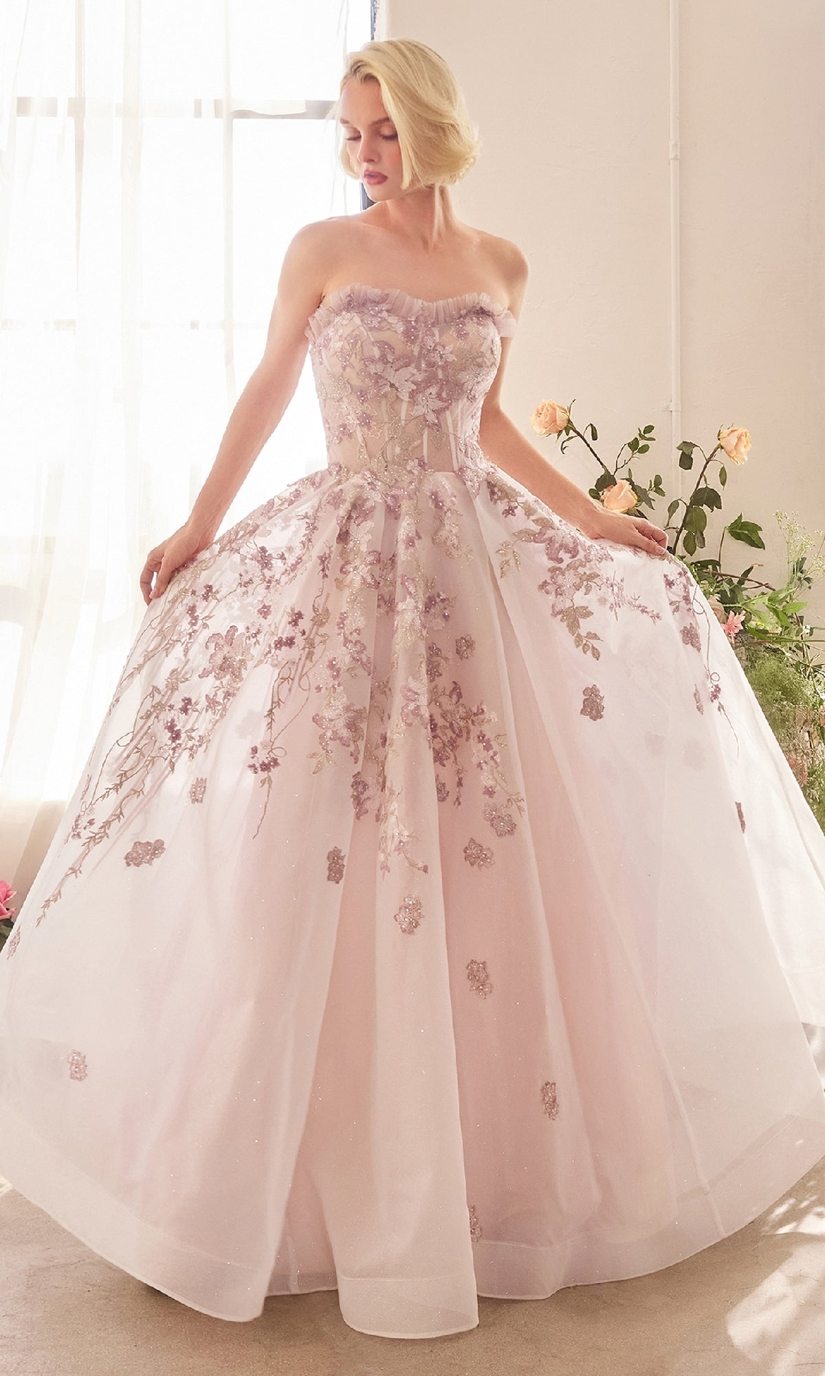 A woman stands indoors wearing the Andrea and Leo A1455 long prom dress, a strapless pastel pink gown with floral lace embroidery and a full tulle skirt.